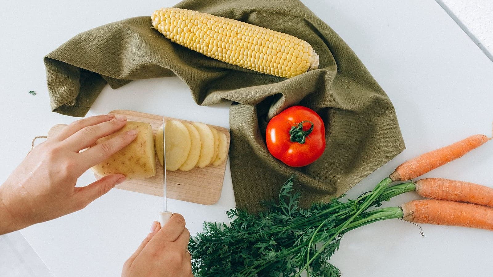 Hands slicing a potato beside fresh carrots and a tomato on a counter.