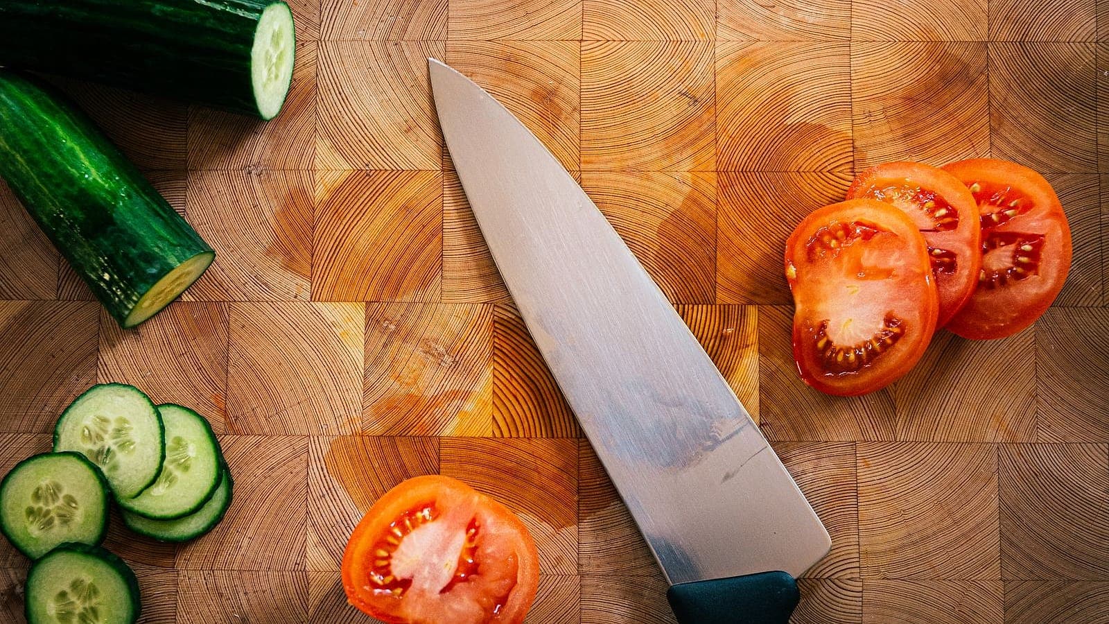 A chef's knife on an end-grain wooden cutting board beside freshly sliced tomatoes and cucumber