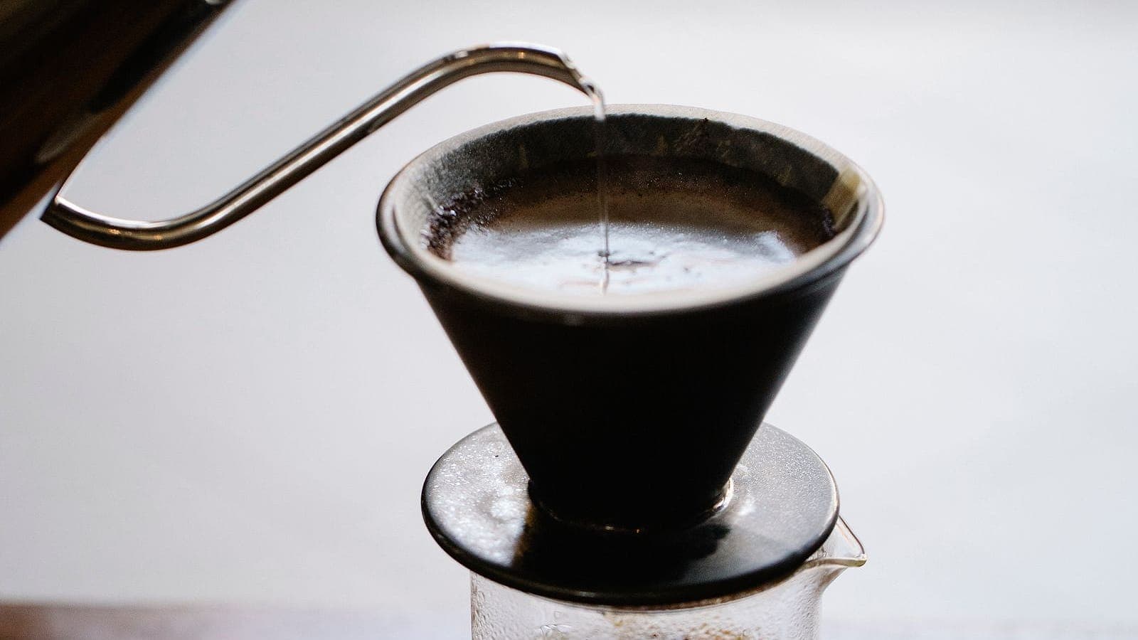 A gooseneck kettle pouring hot water into a cone dripper over a glass beaker.