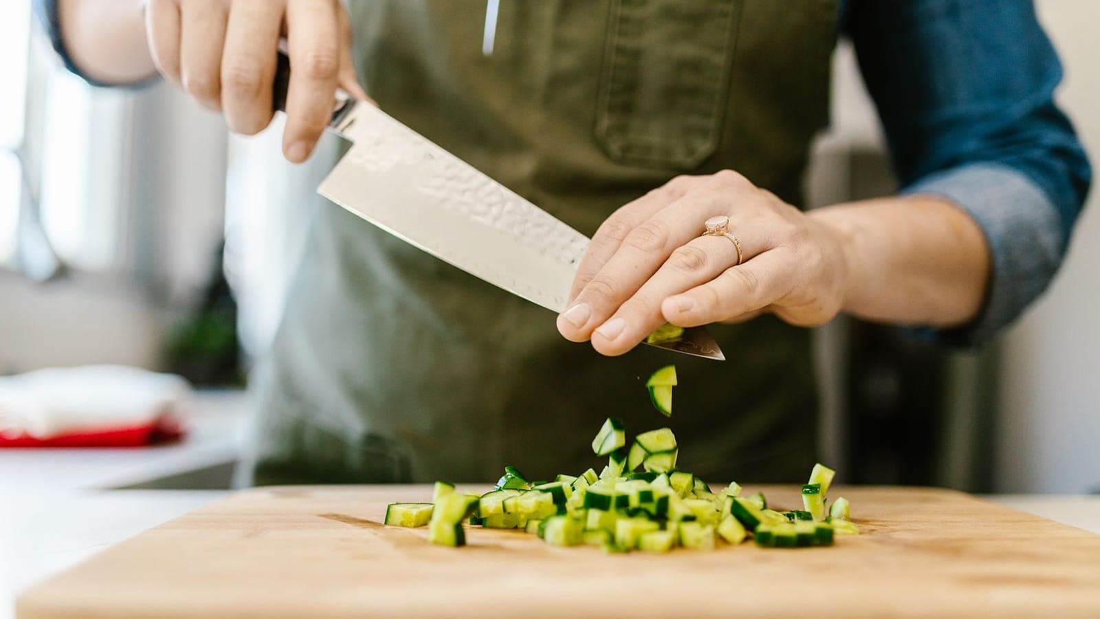 Hands chopping zucchini with a knife on a wooden cutting board.