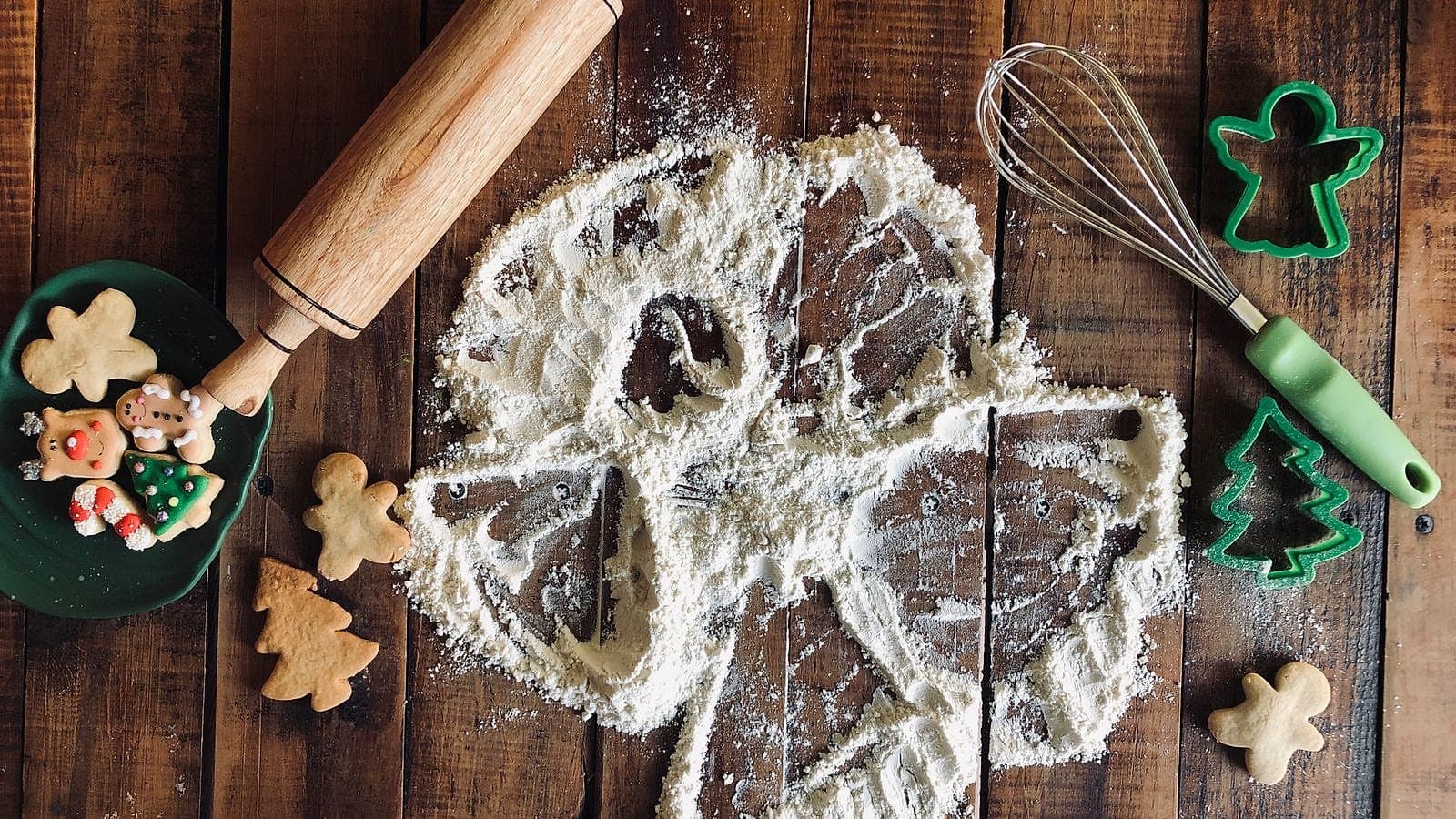Christmas cookie cutters, a rolling pin, and a whisk arranged on flour on a wooden board