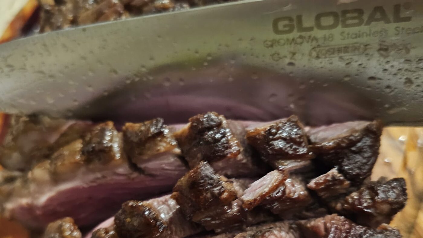 The Global G-2 chef's knife, its CROMOVA 18 etching legible, slicing a cooked picanha steak into medium-rare pieces on a wooden board