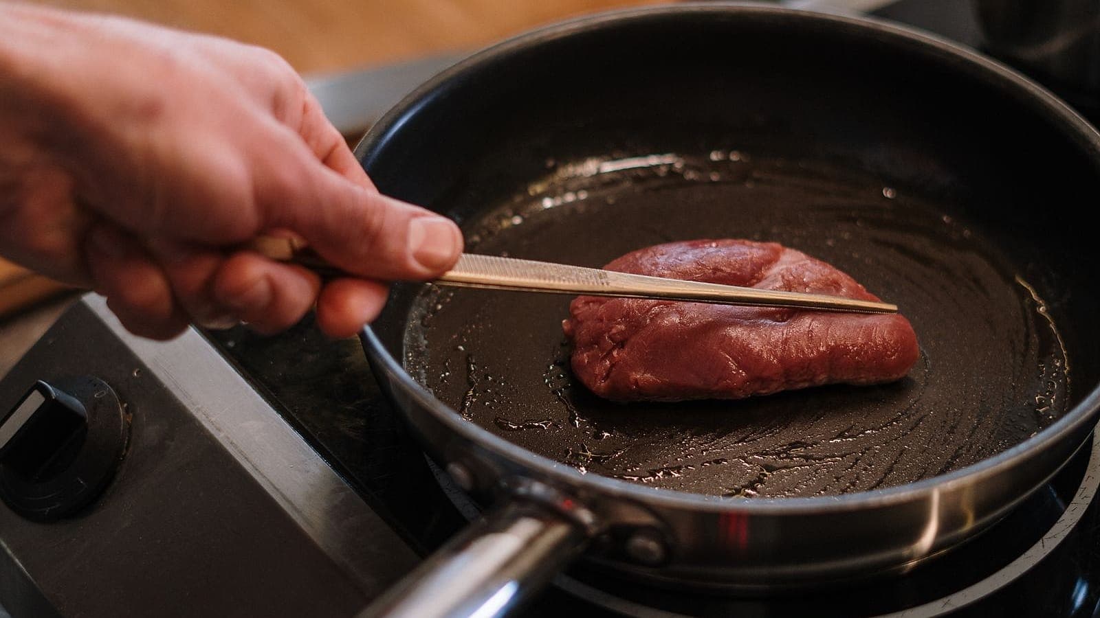 A steak searing in a stainless steel pan, turned with tongs.