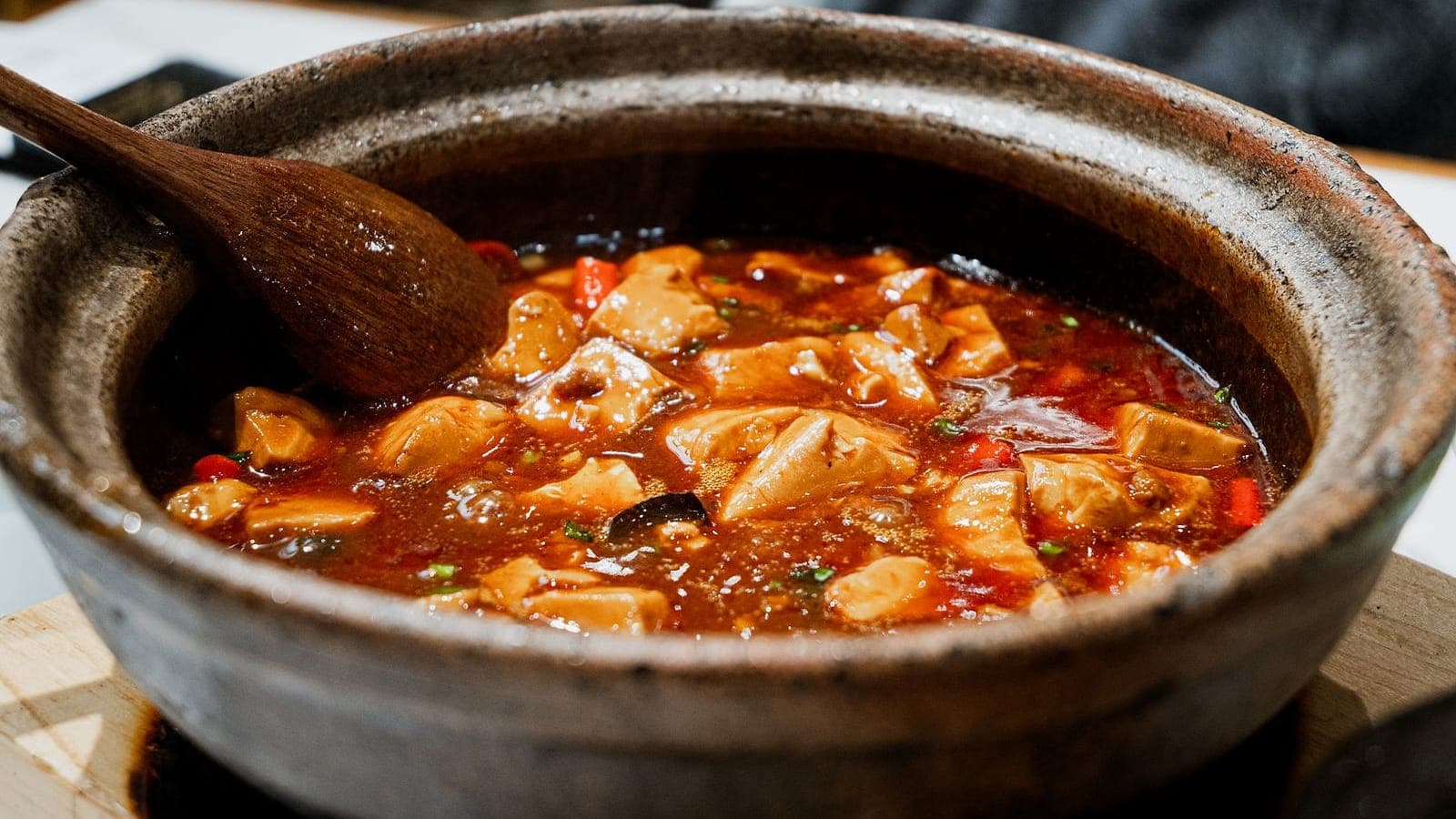 A tofu and vegetable stew in a rustic clay pot with a wooden spoon.